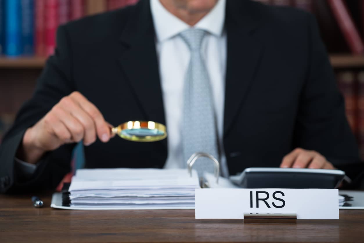 Auditor with a magnifying glass, sitting at a desk marked IRS, examining documents.